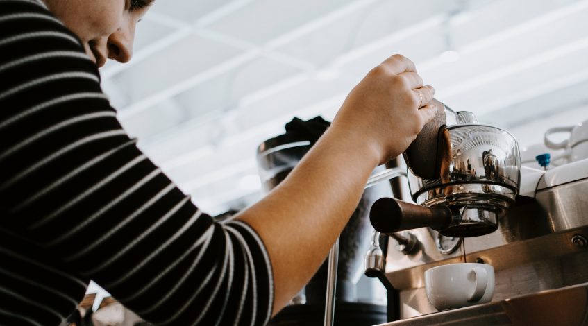 Image of a woman holding down a espresso machine, pouring coffee into the cup.